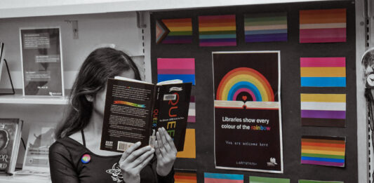 This is a photo of a student in the libarary. There are rows of bookshelves behind them, and they are holding a book titled, “Queer: The Ultimate LGBTQ Guide for Teens.” They are also standing in front of a rainbow poster which reads, “Libraries show every colour of the rainbow, you are welcome here.”