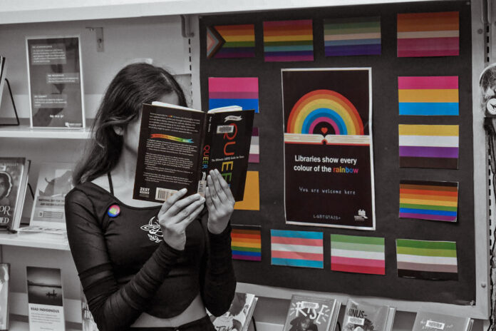 sogi This is a photo of a student in the libarary. There are rows of bookshelves behind them, and they are holding a book titled, “Queer: The Ultimate LGBTQ Guide for Teens.” They are also standing in front of a rainbow poster which reads, “Libraries show every colour of the rainbow, you are welcome here.”