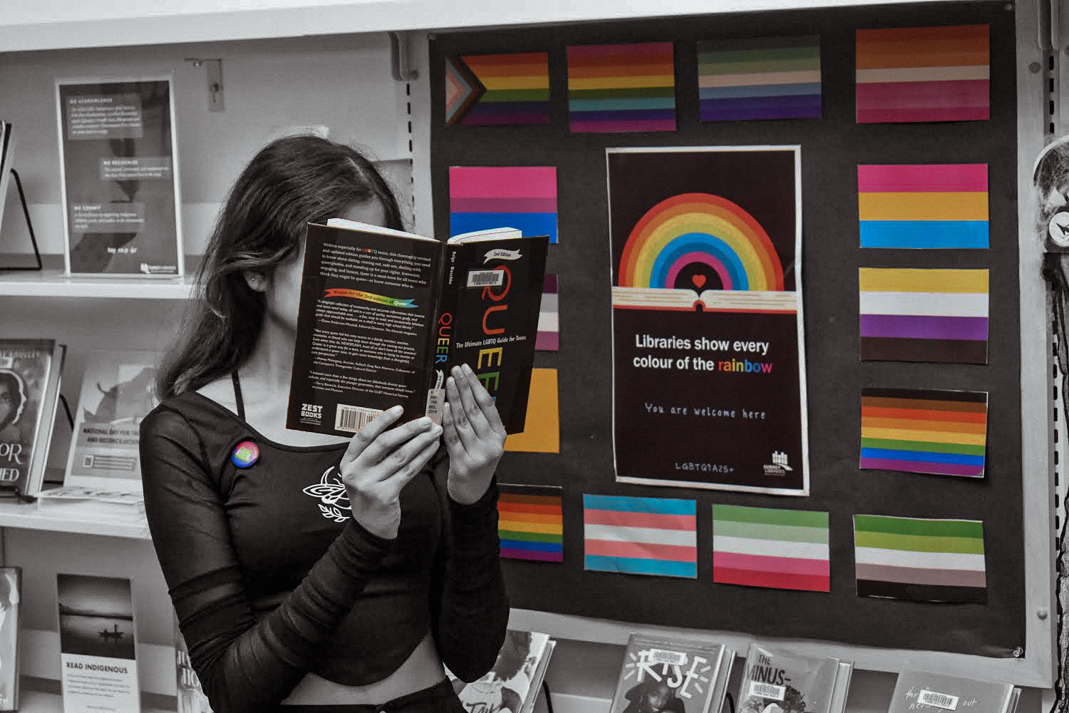 This is a photo of a student in the libarary. There are rows of bookshelves behind them, and they are holding a book titled, “Queer: The Ultimate LGBTQ Guide for Teens.” They are also standing in front of a rainbow poster which reads, “Libraries show every colour of the rainbow, you are welcome here.”