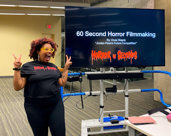 vpl-horror Photo of Doaa Magdy, a Black woman with a red afro wearing a T-shirt that reads “horror in seconds” in a red, gothic font. She is smiling wide with peace hand signs. She is standing in front of a TV screen that reads “60 second horror filmmaking, by Doaa Magdy, Jordan Peele’s Future Competitition.