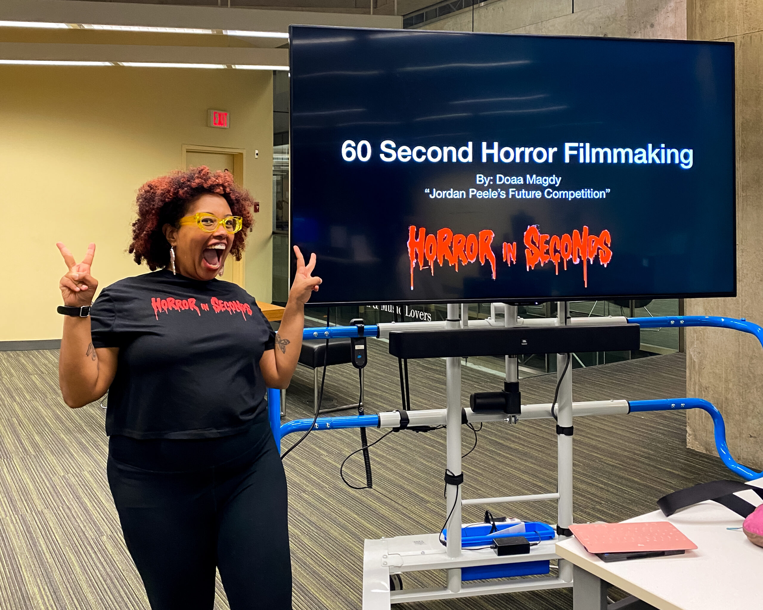 Photo of Doaa Magdy, a Black woman with a red afro wearing a T-shirt that reads “horror in seconds” in a red, gothic font. She is smiling wide with peace hand signs. She is standing in front of a TV screen that reads “60 second horror filmmaking, by Doaa Magdy, Jordan Peele’s Future Competitition.