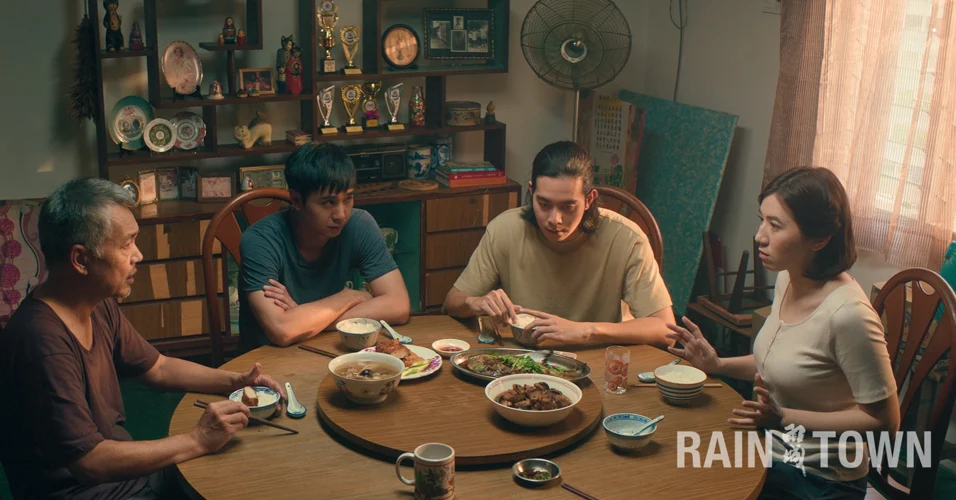 A Cantonese family of four sitting at a dining table with various hot dishes, and bowls of rice. The daughter and father look at each other in a serious way, while the two sons look away with similar expressions. Behind them is a display shelf with many trophies and medals.