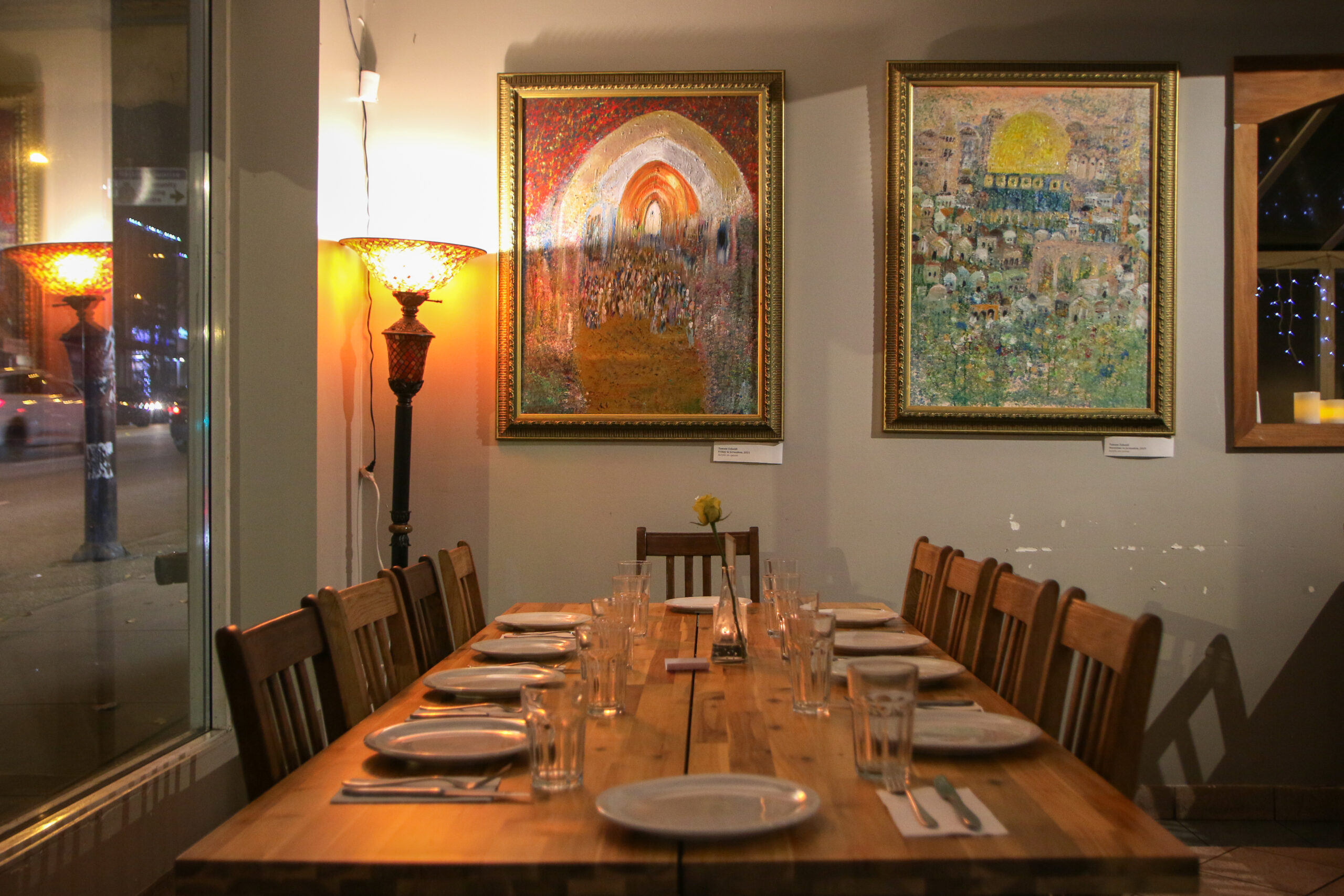 A long wooden table set with empty dishes in front of a moody amber light and acrylic paintings with golden frames on the wall. The window outside shows it is dark out.