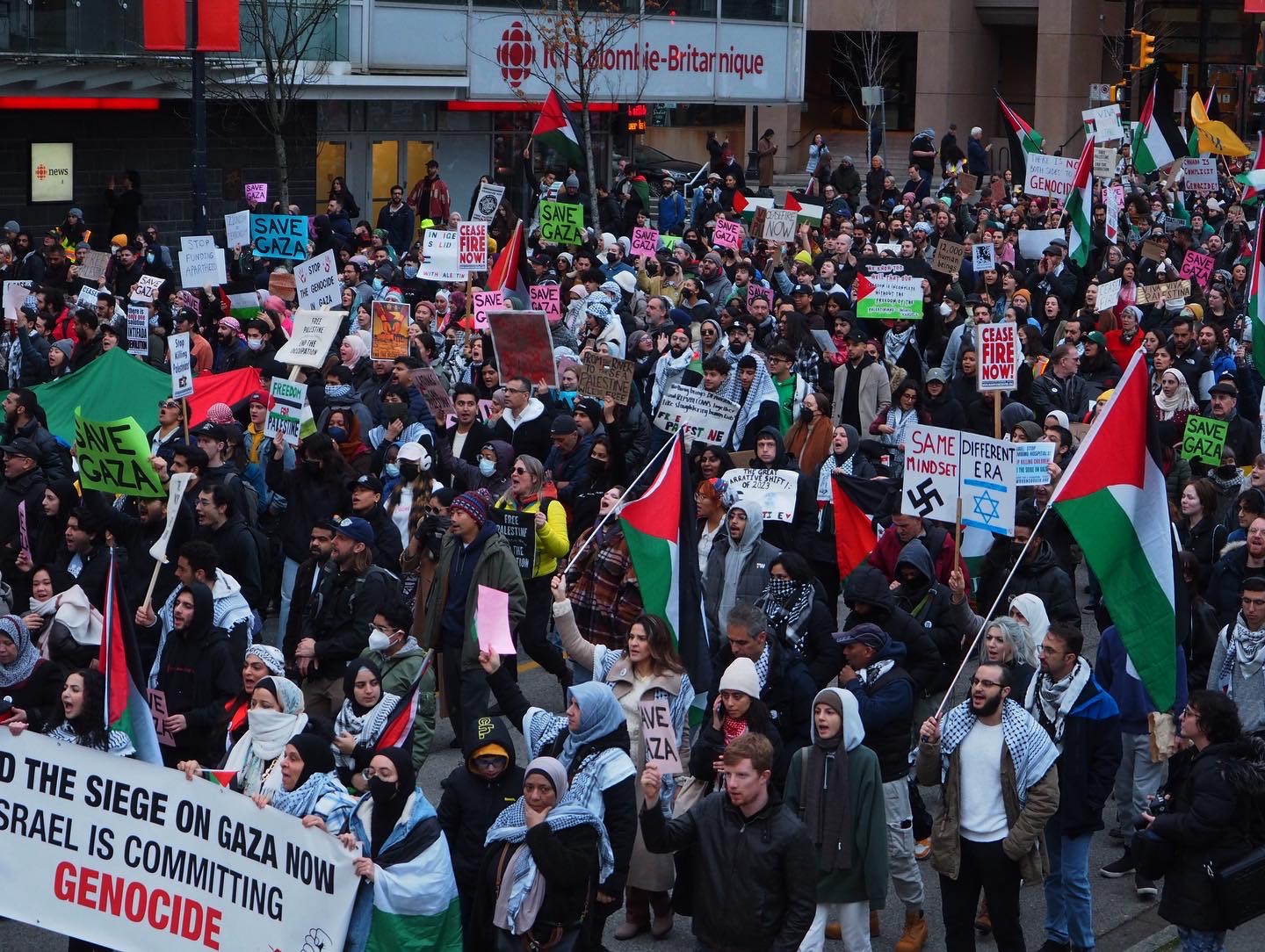 This is a photo of the protests at the Vancouver Art Gallery for Palestine. The crowd of people is walking down the street. They are holding Palestinian flags and signs that say, “Israel is committing genocide,” and “ceasefire now.”