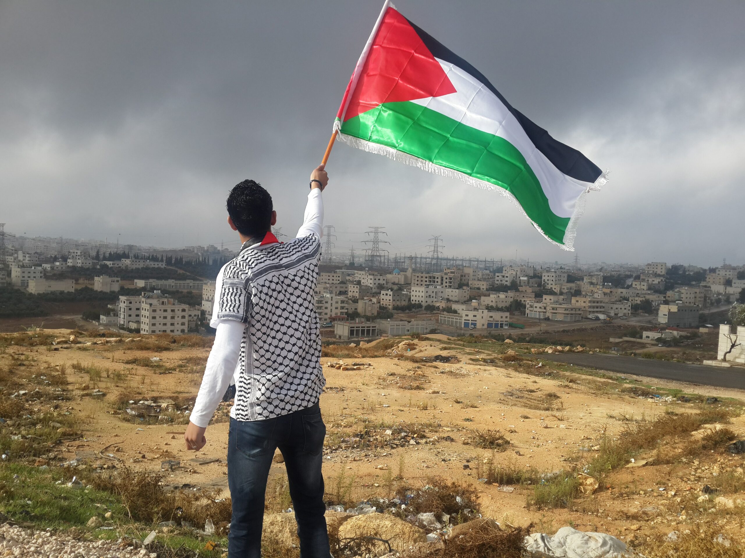 This is a photo of a man standing above a city in Palestine, on a large hill. He is waving a Palestinian flag, red green, and black.