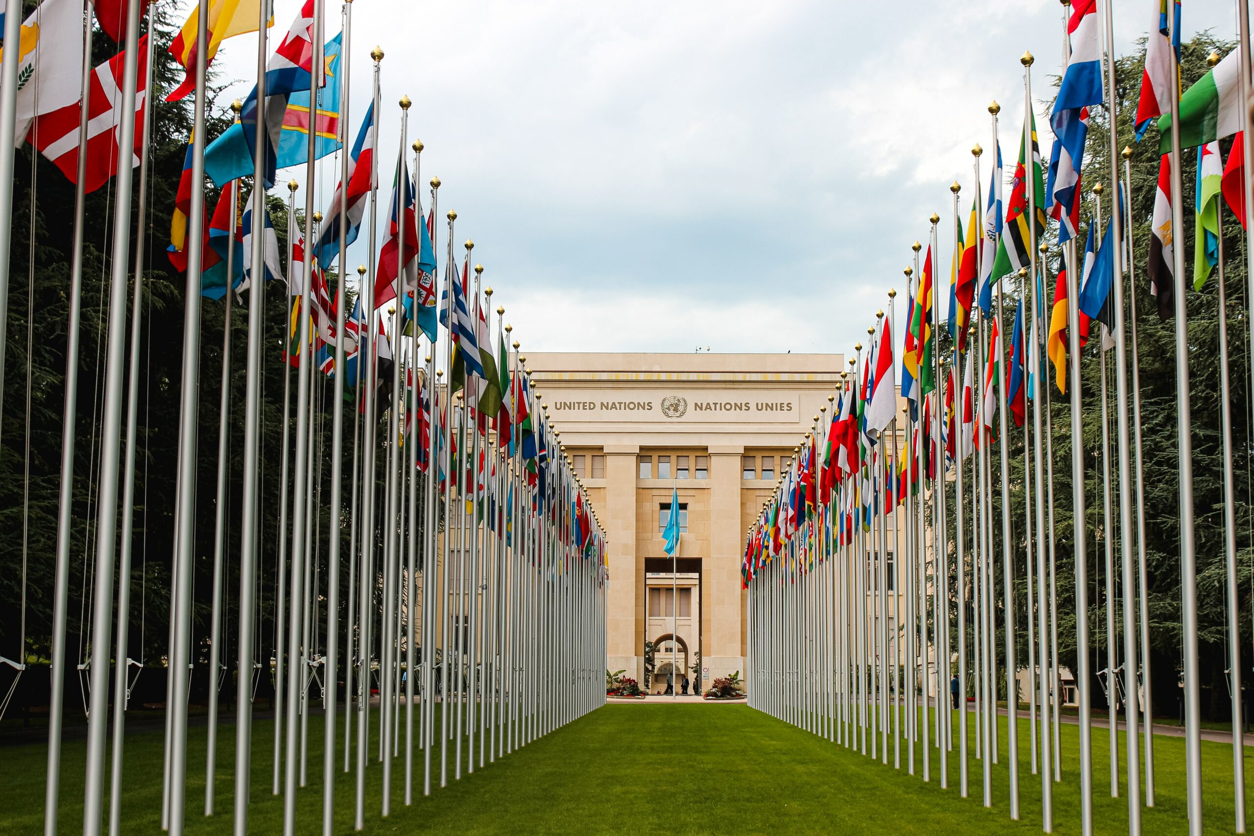 This is a photo of the outside of the United Nations headquarters. Outside the front door is a large row of numerous country flags, lining the grass.