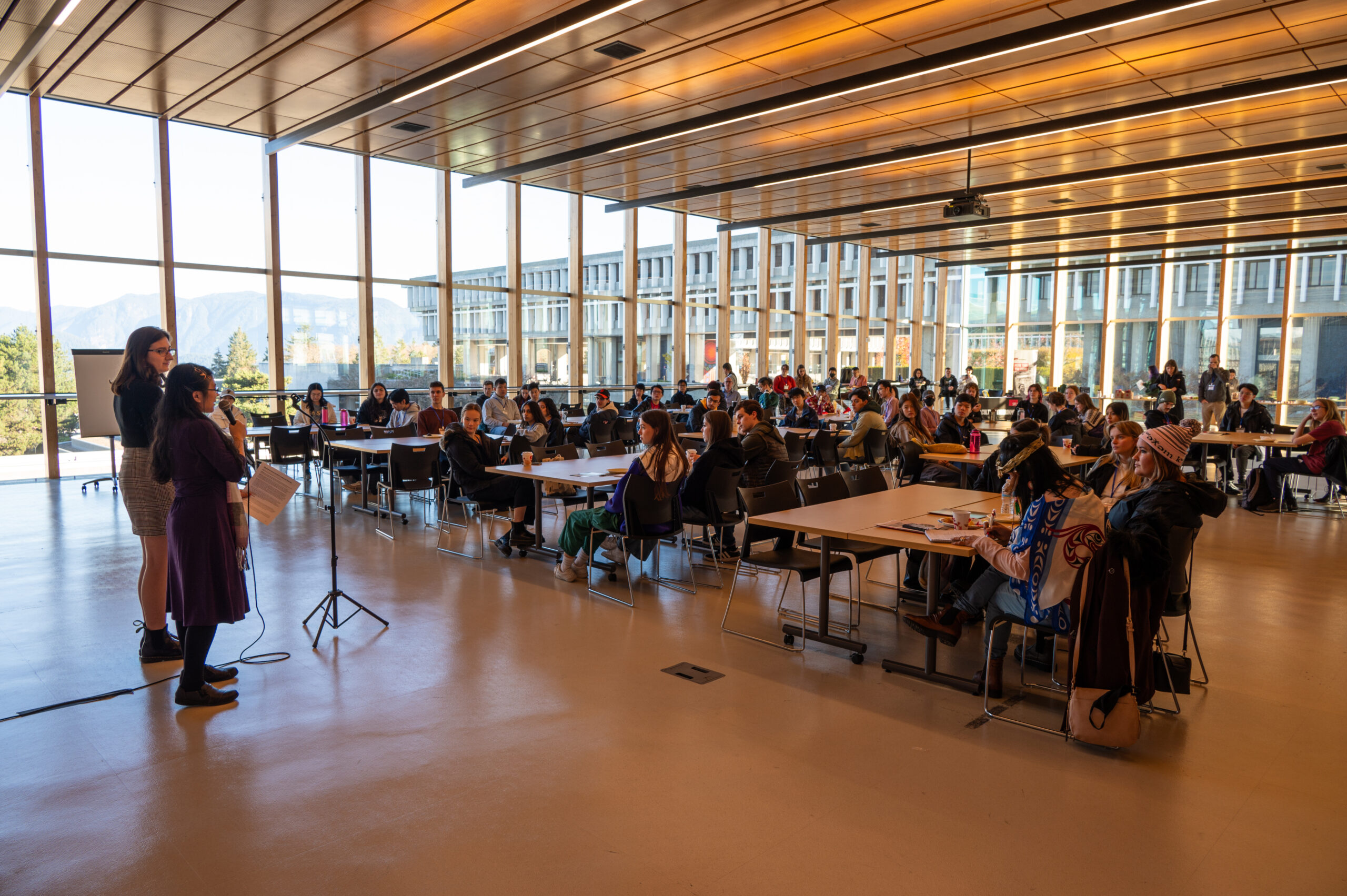 this is a photo of one of the conference presentations. There is a large crowd sitting at tables, looking at the speaker who is at the front of the room, speaking into a microphone