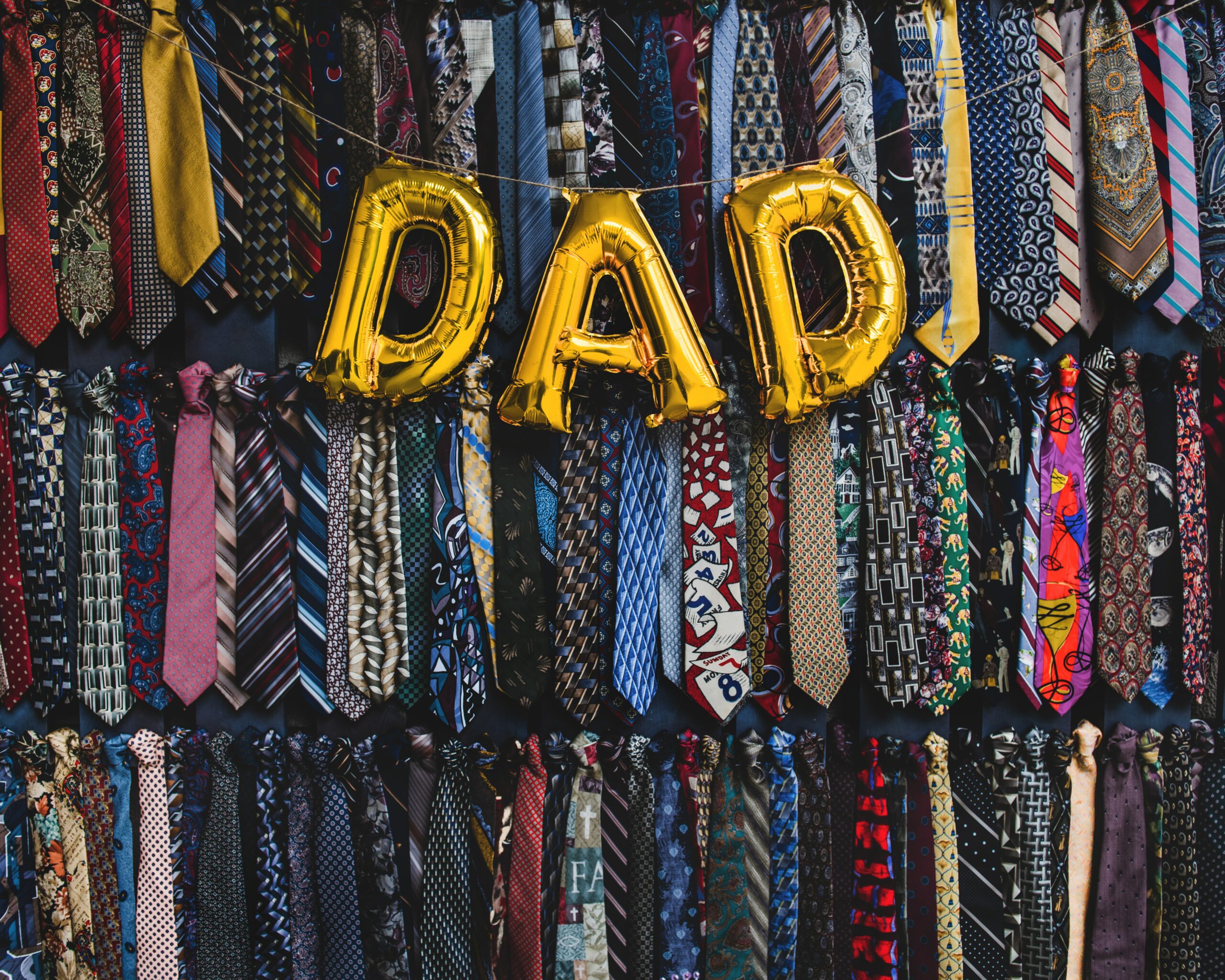 Gold balloons spelling “dad” in front of a wall of ties.