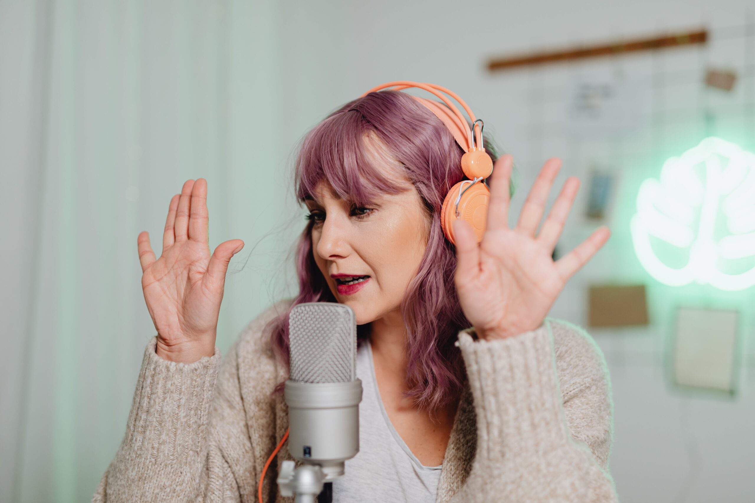 Woman with pink hair and orange headphones talking into a mic.