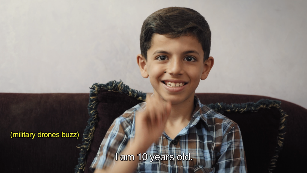 A film still of Vibrations from Gaza. A smiling young boy from Palestine being interviews has his hand up using sign language and caption reads “I am 10 years old.” on the side, text reads “military drones buzzing.”