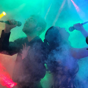 A photo of James Chapeskie and another person singing a duet, each into their own microphones, their silhouettes covered by a fog of coloured lights.