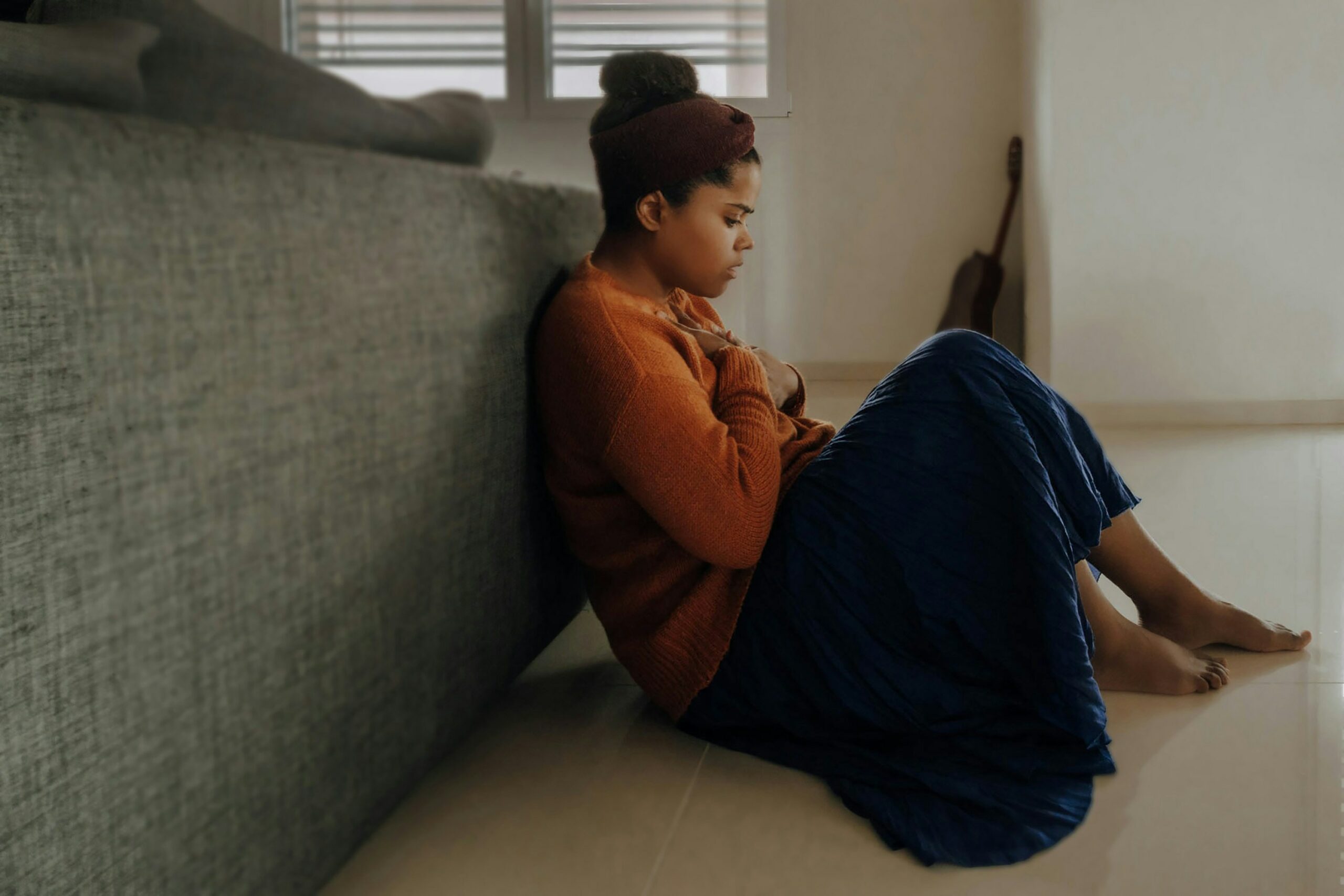 This is a photo of a young person sitting on the floor, leaning against the back of their couch. They look upset, with their head resting in their hands.