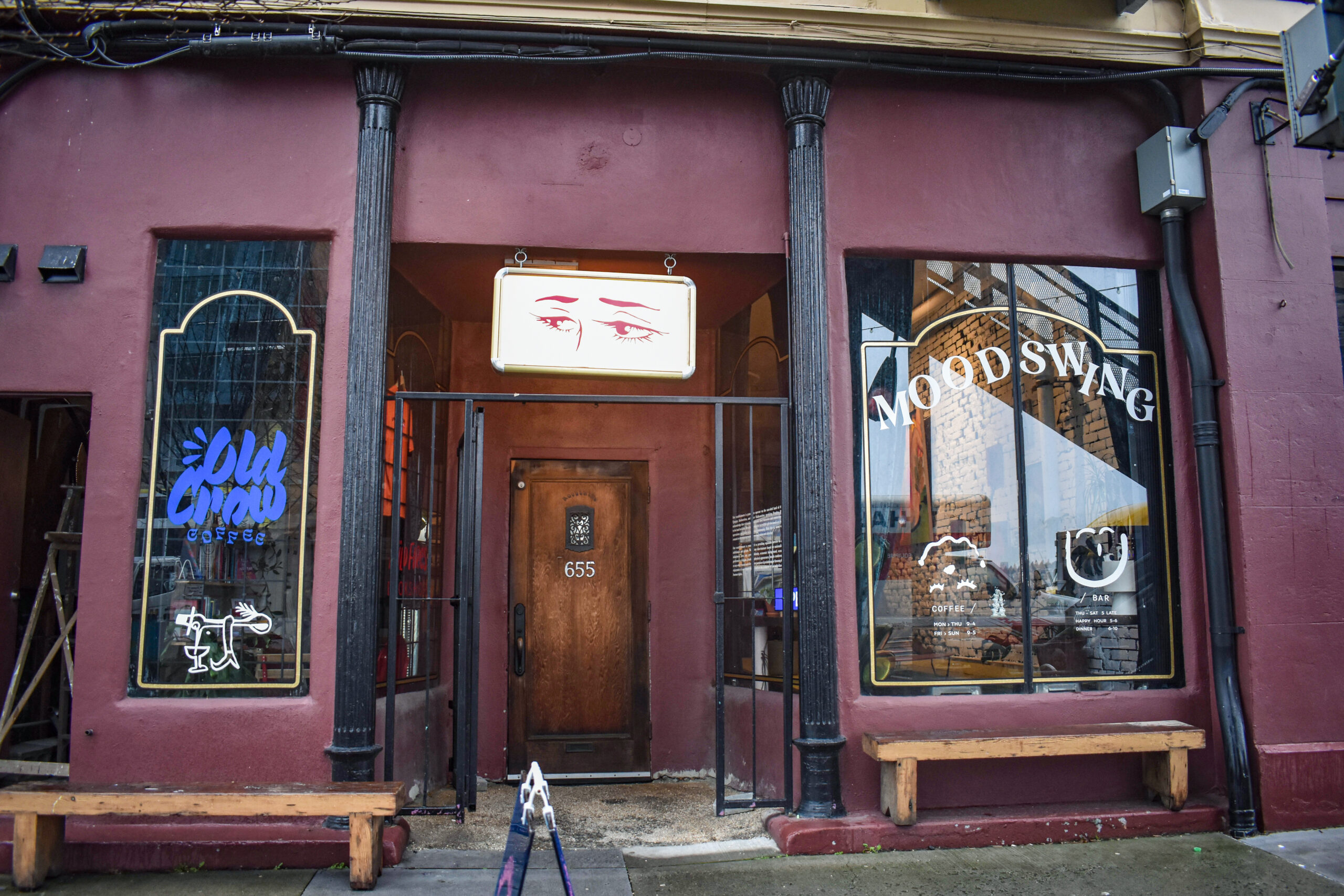 A photo of the exterior of the Moodswing cafe building. The building has an antique appearance with decorative black pillars on either side of the door, carmine and chesnut paint, and two big windows with the words “moodswing cafe” pasted on one and “old crow coffee on another.” Above the door is a poster of a drawing of a set of seductive eyes in maroon ink.