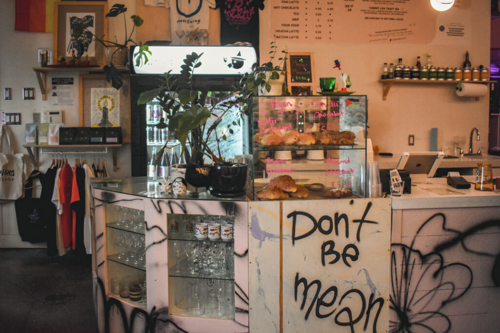 An ordering counter inside moodswing cafe. Behind it are shirts and tote bags on a rack. There is a plant and pastries on display. The front of the counter is a light pink and has graffiti on it and the words “Don’t be mean” are written.