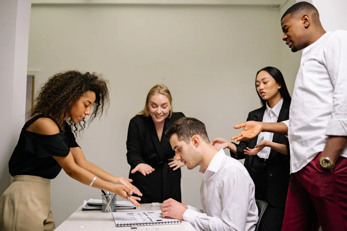 A group of work colleagues gathered over their coworker, who looks distressed starting at their laptop.