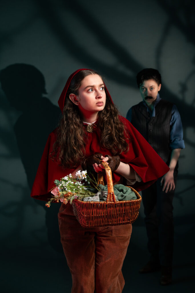 An actor from Into the Woods dressed as Little Red Riding hood holds a basket full of flowers and looks into the distance while a person in the shadows behind her looks at her. The shadows are of barbed wire.
