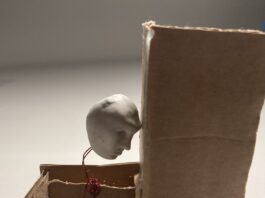 An automata of a white sculpted head, wired to a cardboard handle, banging against a wall.
