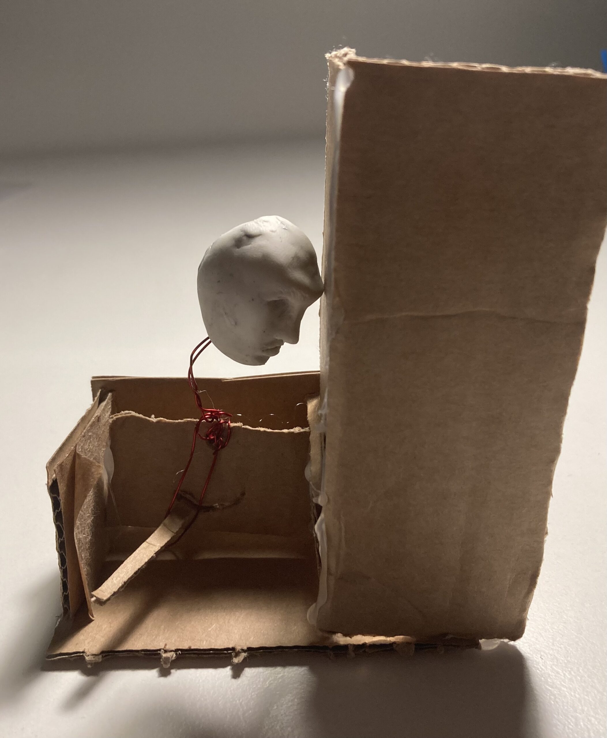 An automata of a white sculpted head, wired to a cardboard handle, banging against a wall.