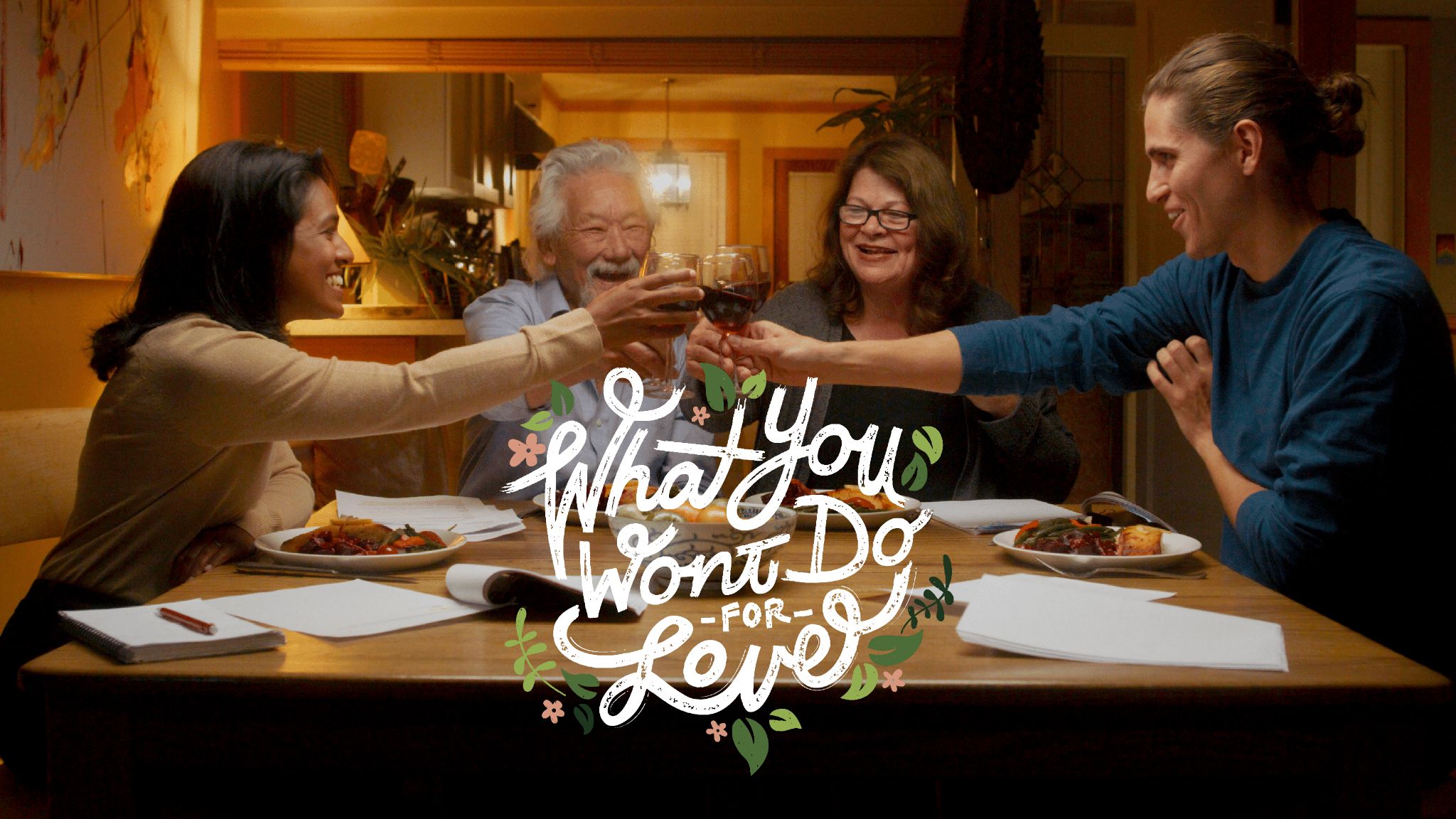 Miriam Fernandes, David Suzuki, Tara Cullis, and Sturla Alvsvaag sit around a dinner table scattered with scripts and food. They raise their wine glasses to the centre in cheers. Underneath is the play’s logo, “What You Won’t Do For Love” in white cursive surrounded by green leaves and pink flowers.