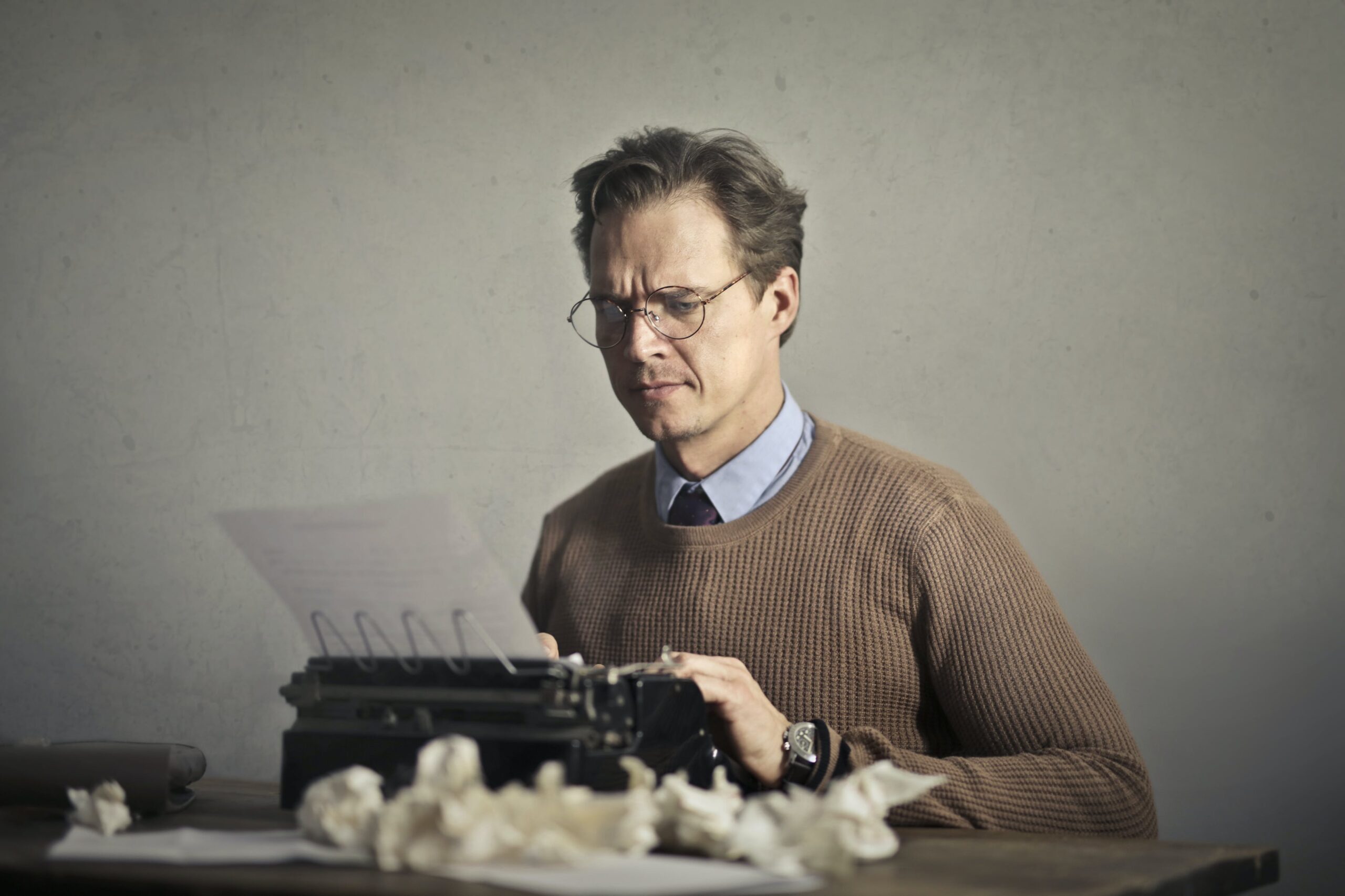 A man wearing a knit sweater furrows his brows, typing furiously on a typewriter.