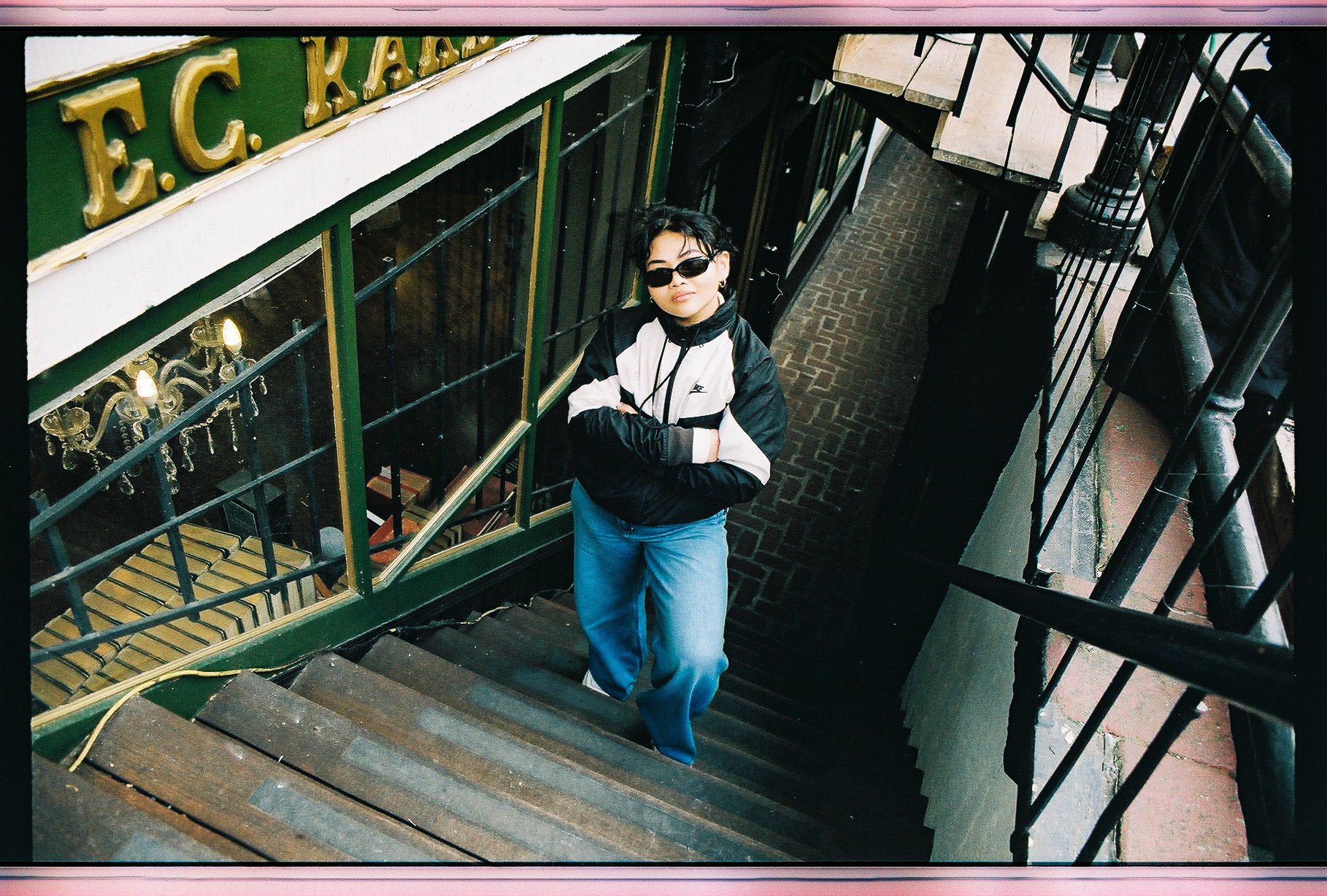 Etchos posing in a black and white bomber jacket, her haid pulled up, and statement sunglasses on. She’s standing at the bottom of the stairs to an antique shop.