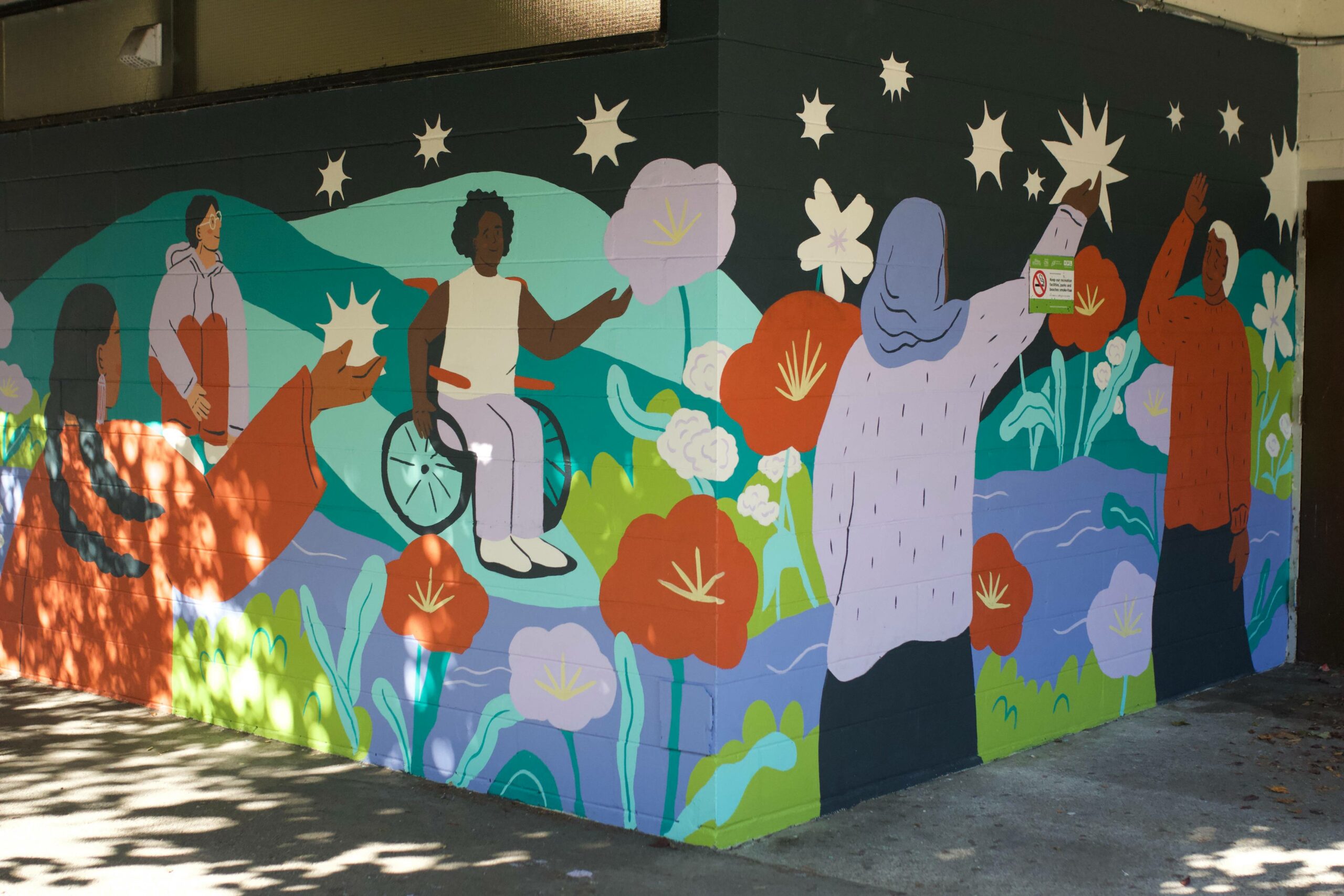 “Hate to Hope” a heartwarming mural of an elderly woman looking upon the younger generation, reaching for and catching stars — a symbol of hope. It invites eyes to a vivid mountainscape and clusters of local flowers meant to represent “growth, endurance, and peace,” that seem to sprout right from the wall. An opaque night sky contrasts with pastel landscapes and clothing.