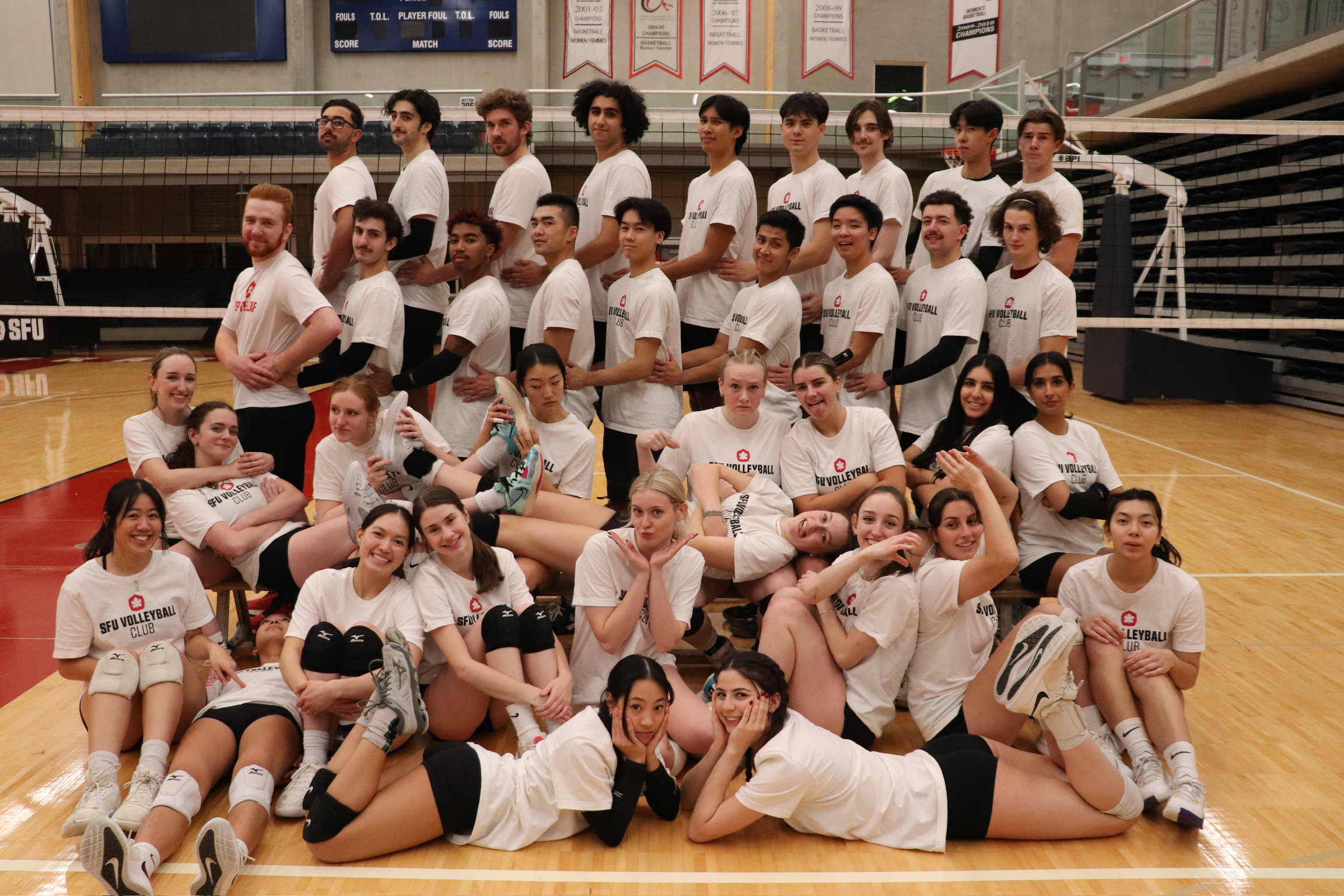 team photo of the SFU volleyball club posed in rows in front of the volleyball net