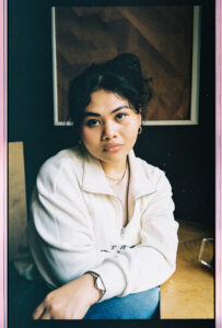 A closeup portrait of Etchos sitting in a cafe looking into the camera with a confident look on her face. Her legs are crossed and she’s wearing a white fleece. 