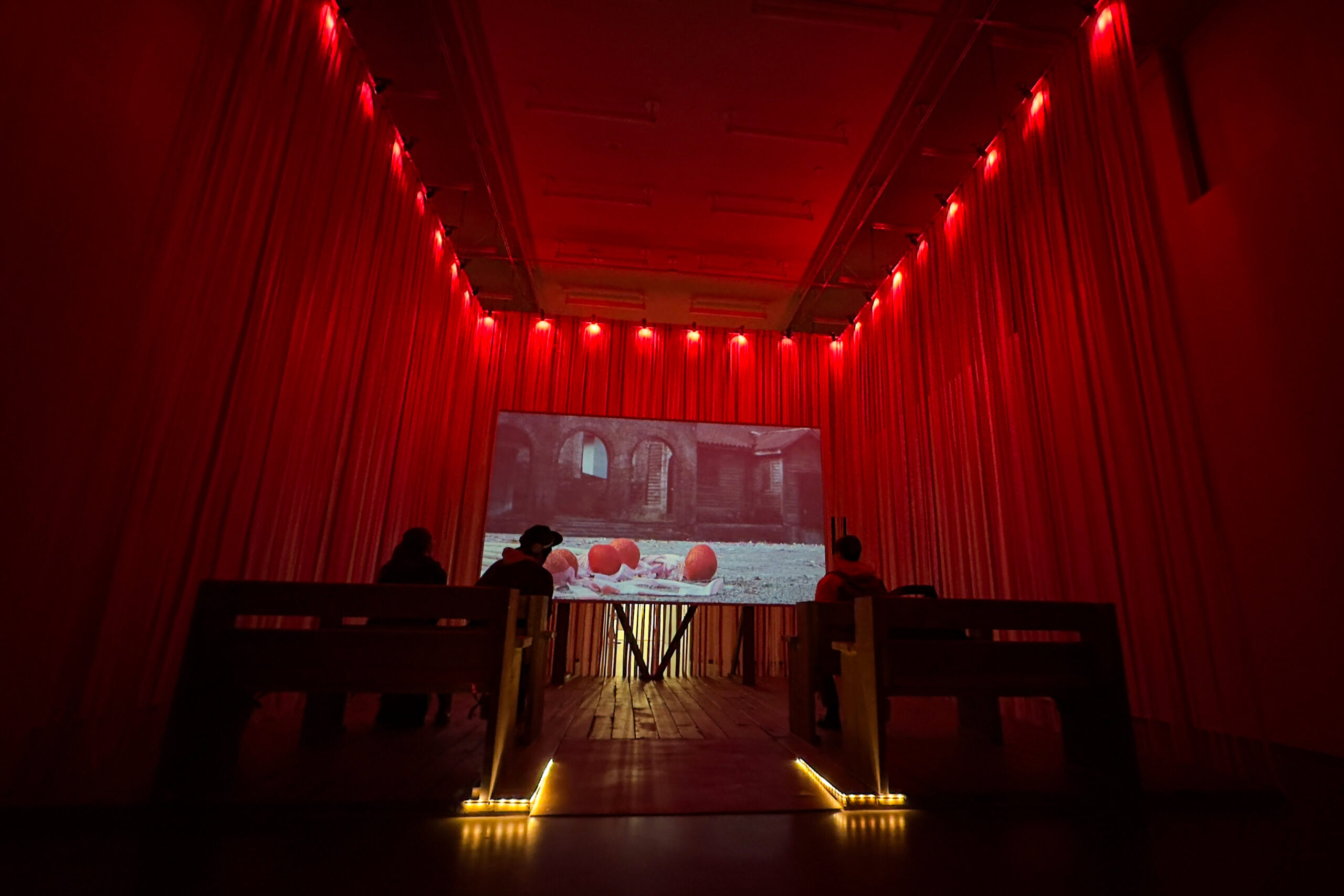 A small room surrounded by red theatre-like curtains and 3 people sitting on benches looking toward a film projected on a screen.