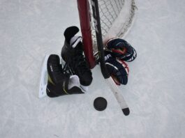 photo of a pair of gloves, skates, and a stick leaned up against a hockey net on an outdoor rink.