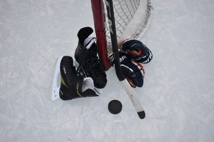 photo of a pair of gloves, skates, and a stick leaned up against a hockey net on an outdoor rink.