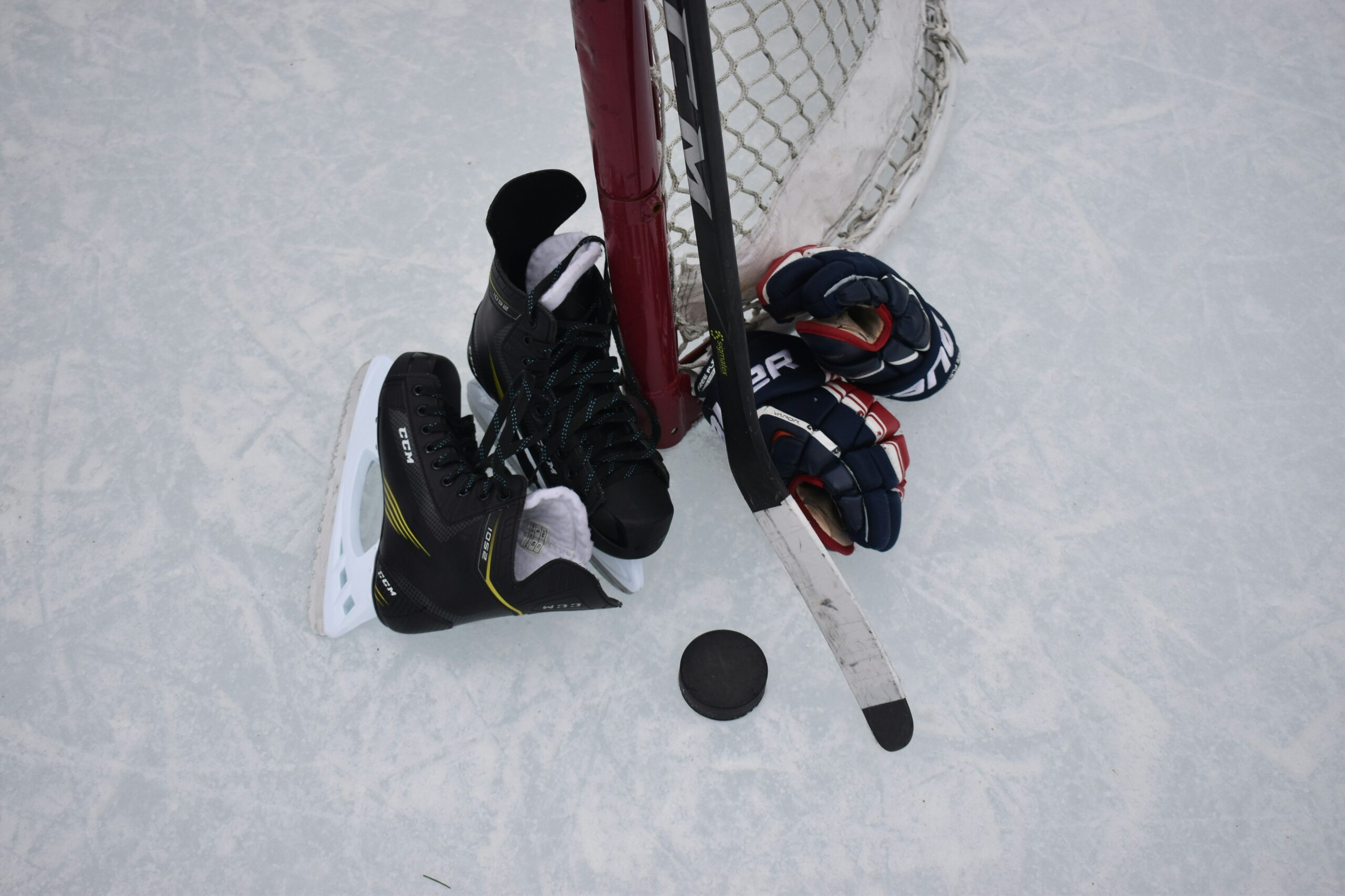 photo of a pair of gloves, skates, and a stick leaned up against a hockey net on an outdoor rink.