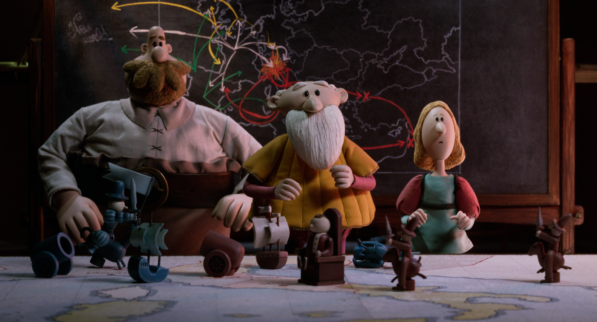 Three clay dolls standing in front of a large map speckled with coloured arrows. On a table in front of them are small figurines including pirate ships, individuals riding on horses, a cannon, and a person sitting on a wooden throne-like chair.