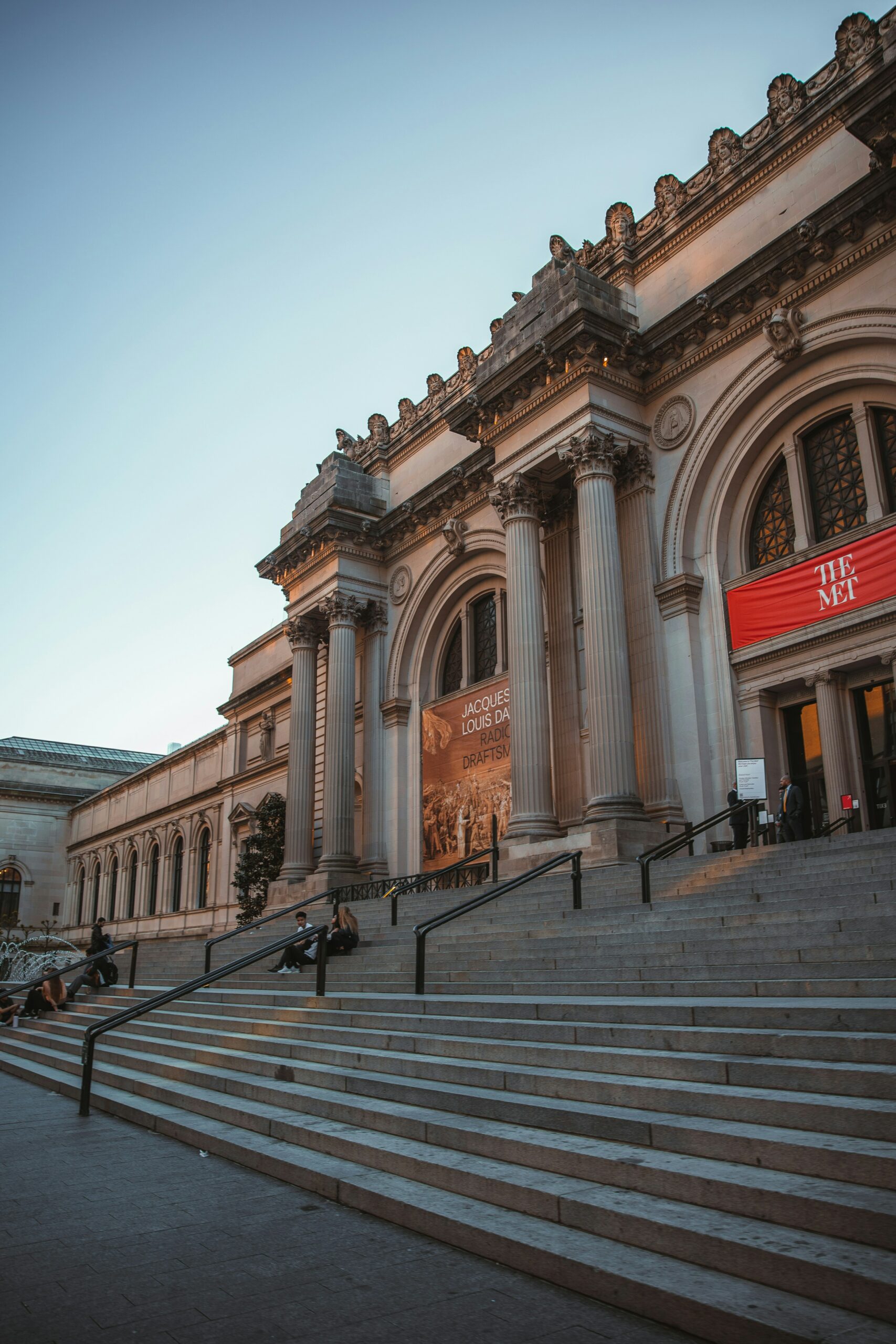 Entrance to the Metropolitan Museum of Art. There is a stone staircase leading up to regal pillars and arched doorways. A banner that reads “The Met” is displayed above the main door.