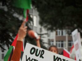 a sign is held up in the middle of a protest, reading “our voices will never be silenced” with an image of the Palestinian flag.