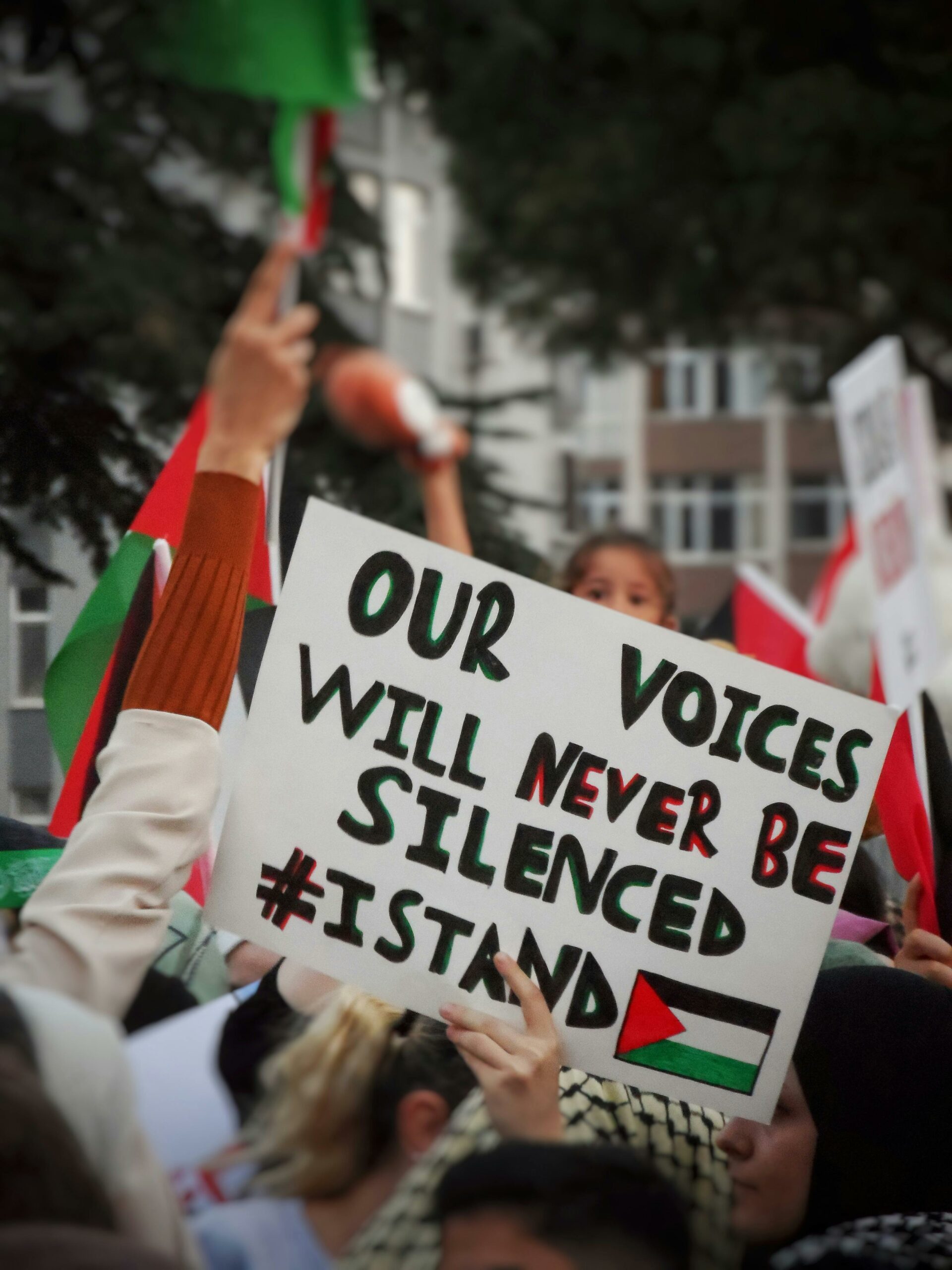 a sign is held up in the middle of a protest, reading “our voices will never be silenced” with an image of the Palestinian flag.