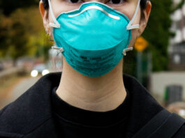 A closeup of a person standing outside wearing a blue N-95 mask covering their nose and mouth.
