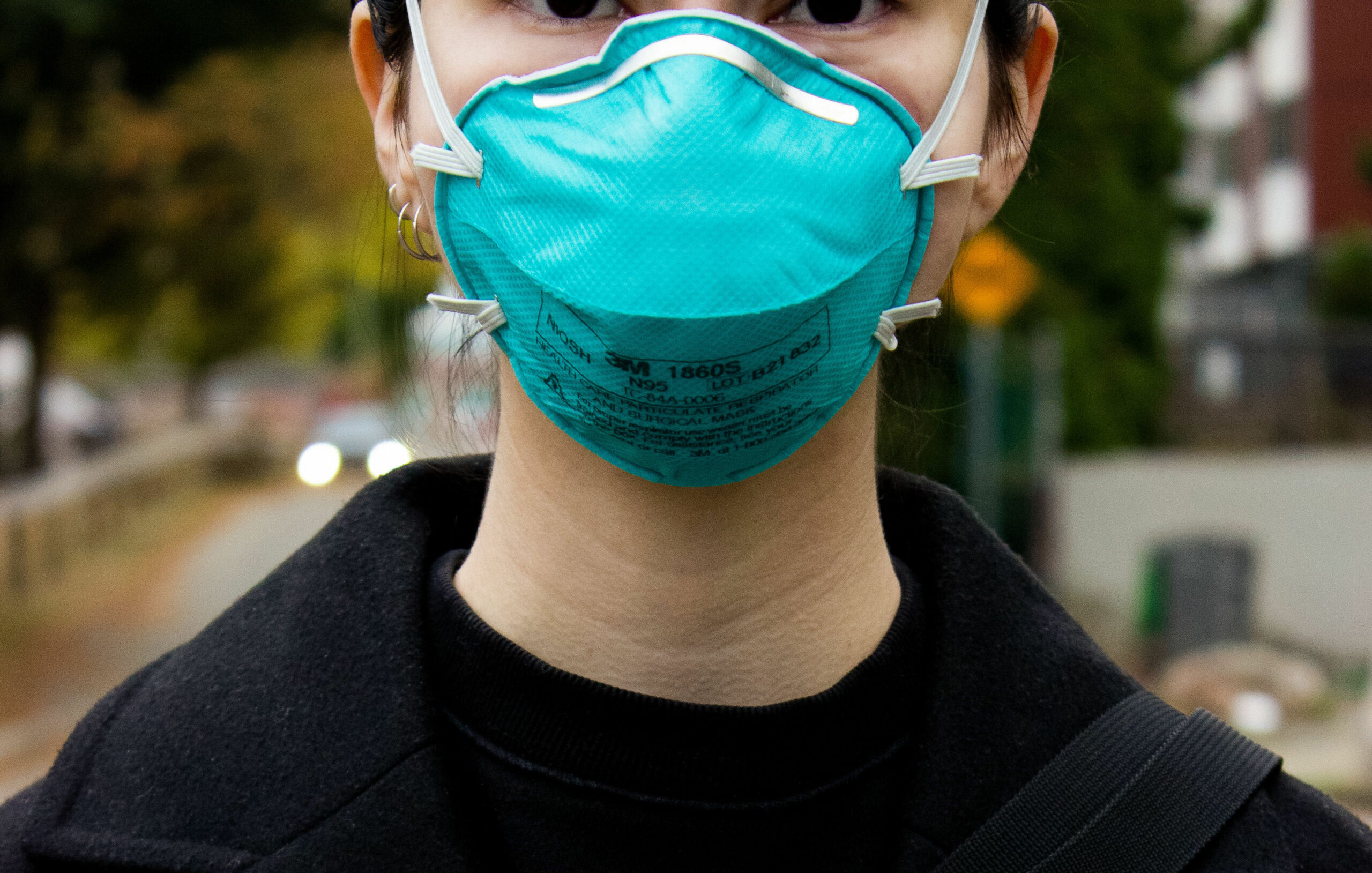 A closeup of a person standing outside wearing a blue N-95 mask covering their nose and mouth.
