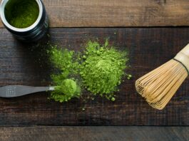 A spoonful of matcha powder on a wooden board alongside a matcha whisk and cup of green tea.