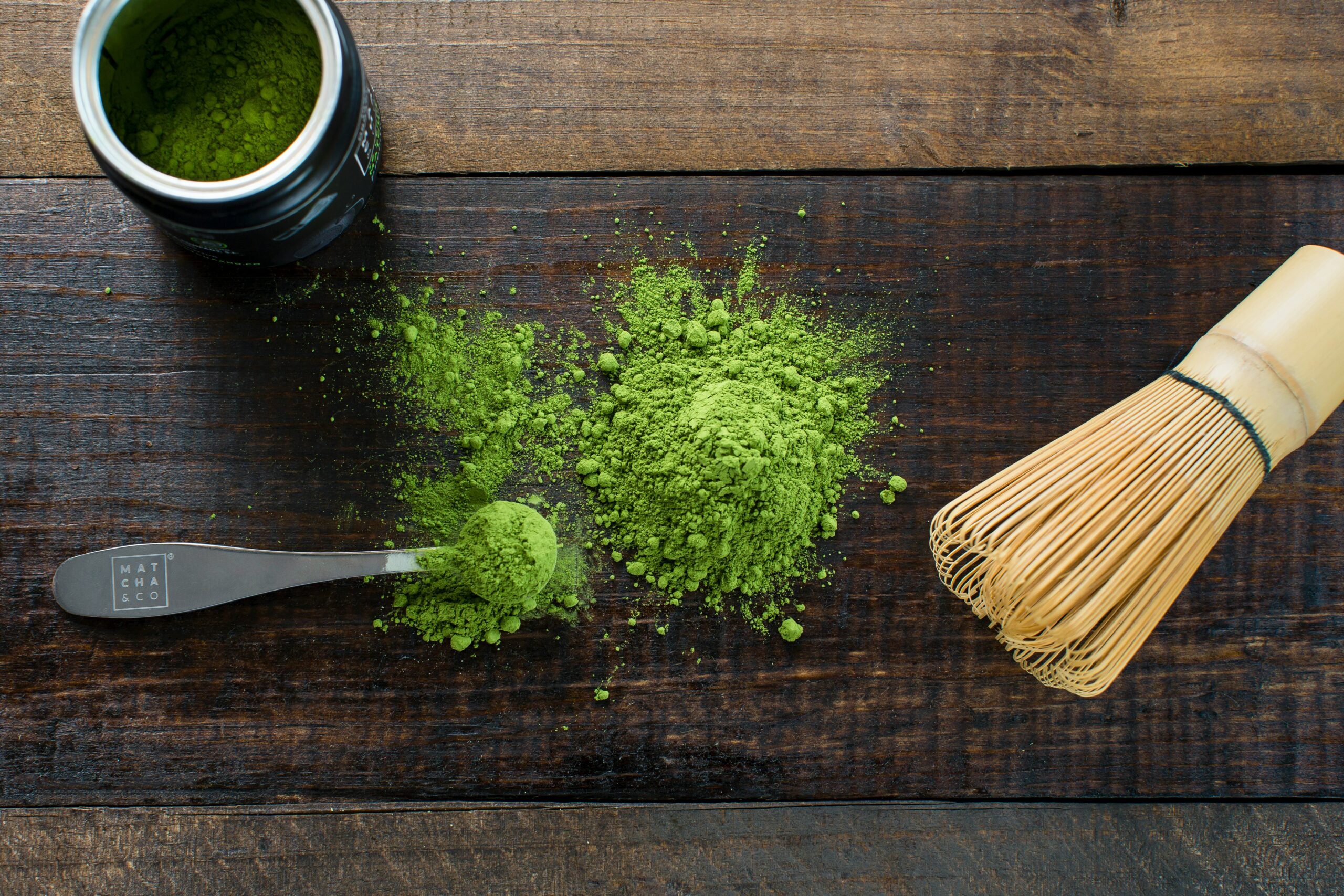 A spoonful of matcha powder on a wooden board alongside a matcha whisk and cup of green tea.