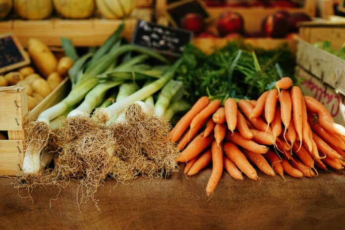 A shelf with vegetables. In focus are green onions and bunches of carrots.