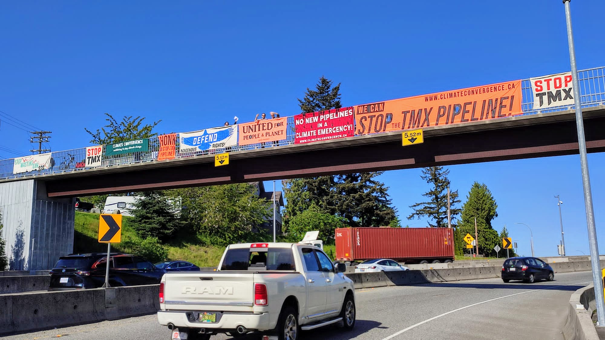 a series of multicoloured banners draped over a freeway. The banners advocate to stop the TMX pipeline, and activists on top of the bridge cheer