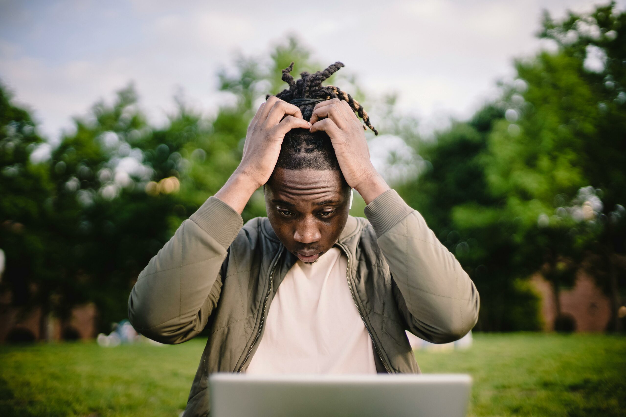 Man sitting outside in front of his laptop. He is stressed and has both his hands on his head.