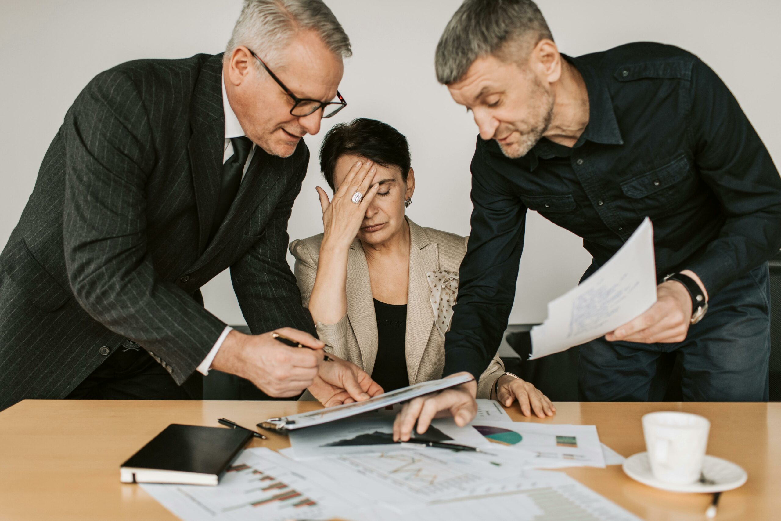 Corporate woman sitting at desk looking overwhelmed by the business papers two men are trying to show her.