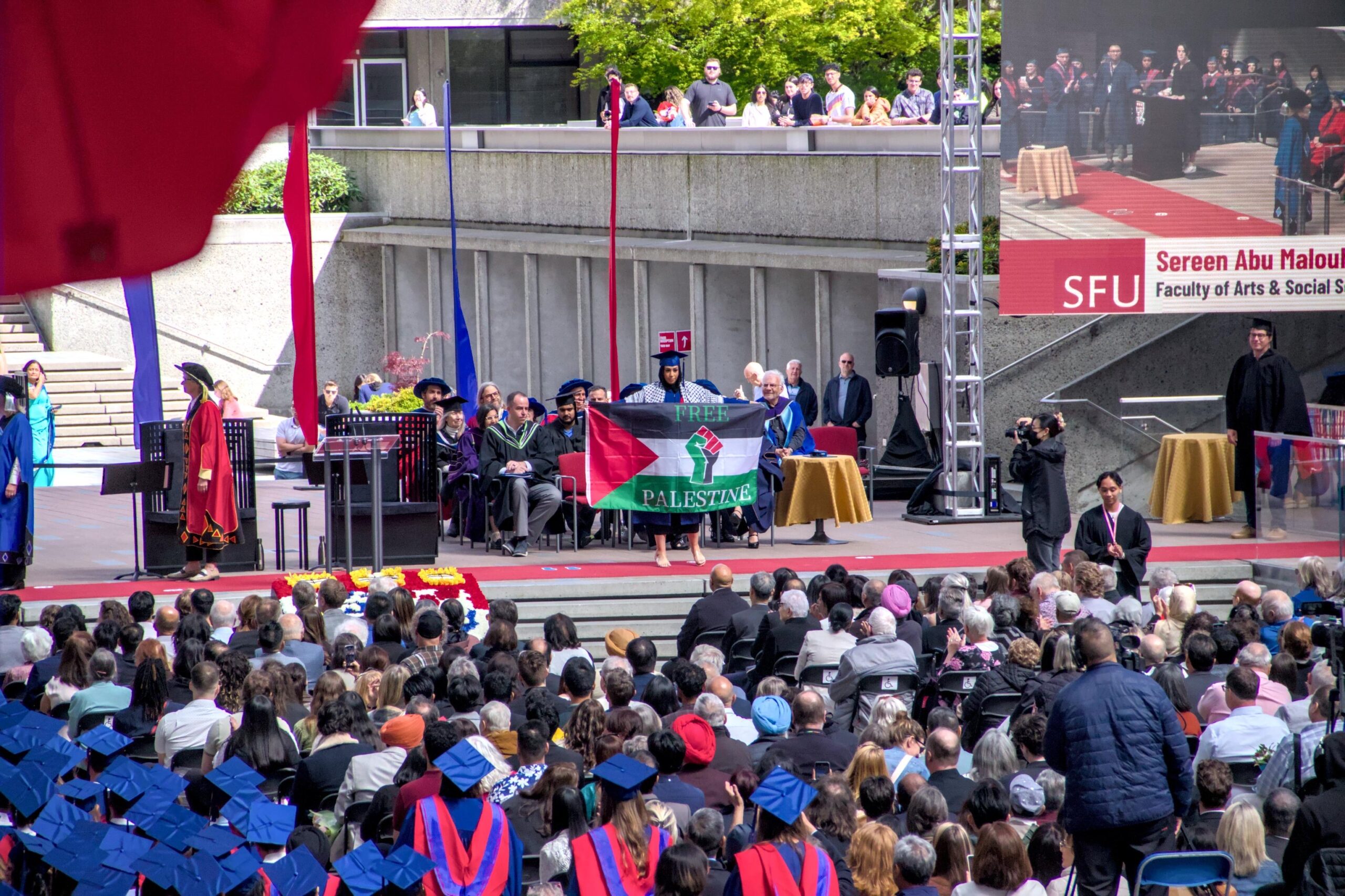 This is a photo of the convocation stage at SFU. As a student crosses the stage, they are holding a large Palestinian flag that reads, “Free Palestine.”