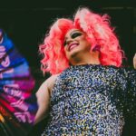 Peakie is here to answer your queeries Drag performer with bright pink hair and a sequined dress holding a fan that says “Girl, bye”