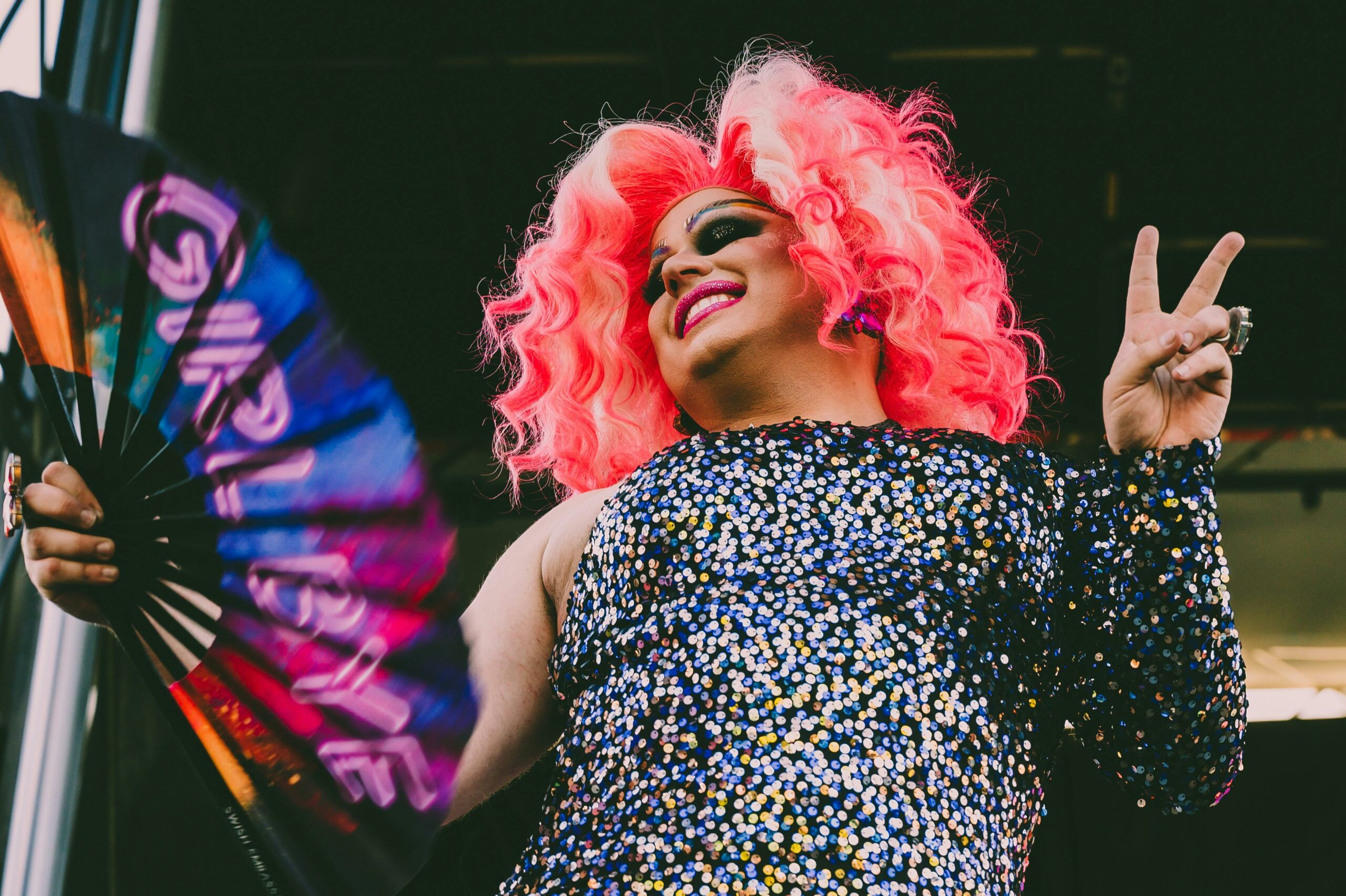 Drag performer with bright pink hair and a sequined dress holding a fan that says “Girl, bye”