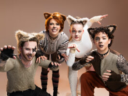 Four individuals wearing cat-like makeup with fluffy cat ears. Two on the outsides stand on one knee, while the two in the middle are slightly crouched. All four have their arms bent and posed as if they are pouncing at the camera.