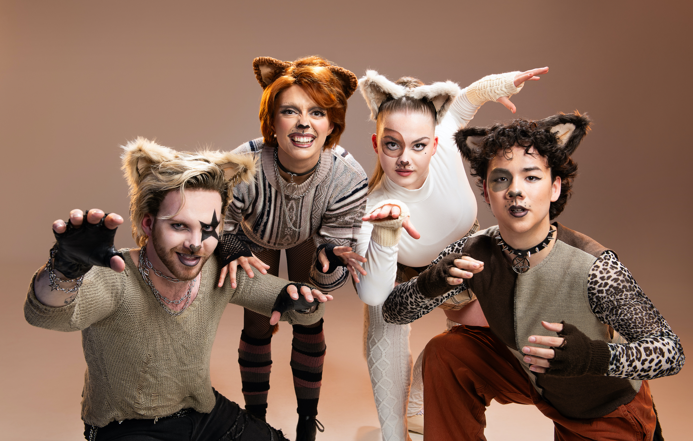Four individuals wearing cat-like makeup with fluffy cat ears. Two on the outsides stand on one knee, while the two in the middle are slightly crouched. All four have their arms bent and posed as if they are pouncing at the camera.