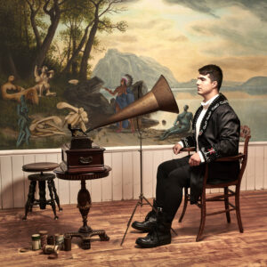 a man sitting at a wooden dining chair while facing a device that looks like a phonograph. Painted on the wall in the background is an image of humanoid shapes and colours interacting with a person wearing an Indigenous headdress. 